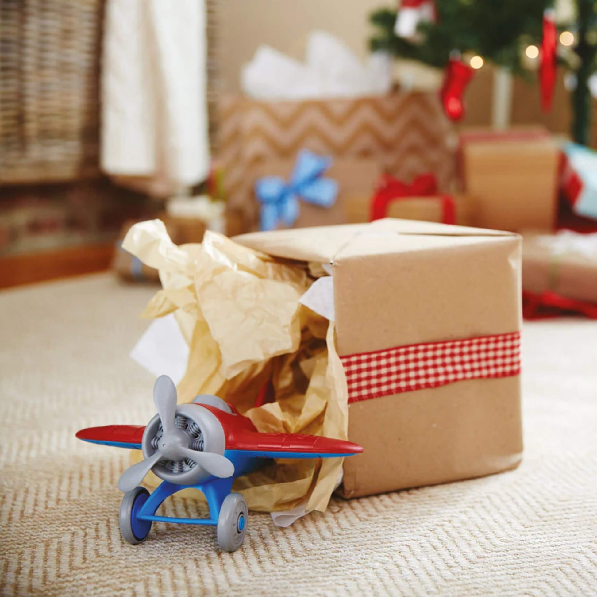 Red toy airplane emerging from a brown paper-wrapped gift box, surrounded by festive Christmas decorations and soft lighting.