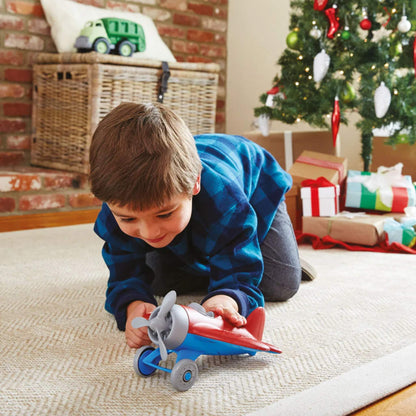 Child in a blue checked shirt playing on the carpet with the red toy airplane in front of a decorated Christmas tree and wrapped presents.