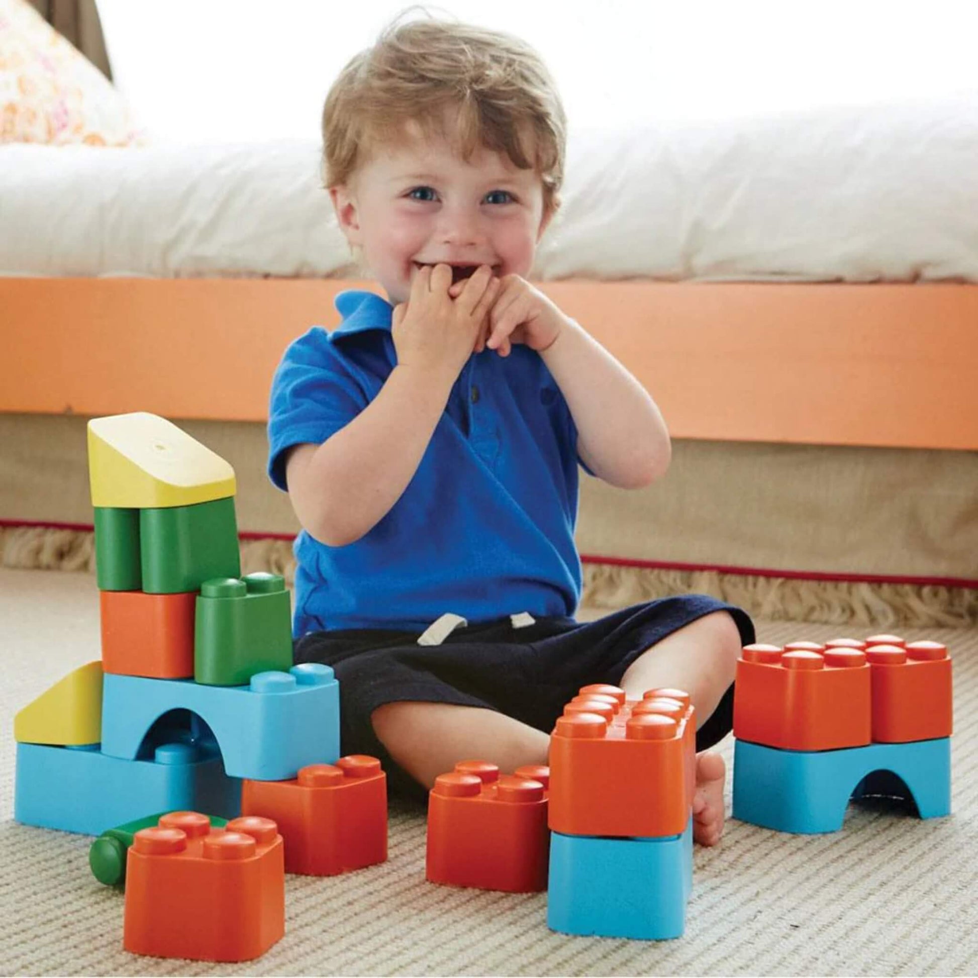 Young child sitting on the floor smiling beside a colourful block tower, surrounded by large interlocking blocks.