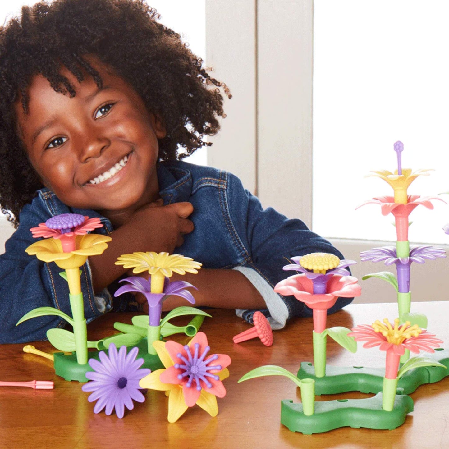 Child smiling at a table surrounded by colourful buildable flowers in pink, yellow, and purple with stems and leaves arranged in green bases.