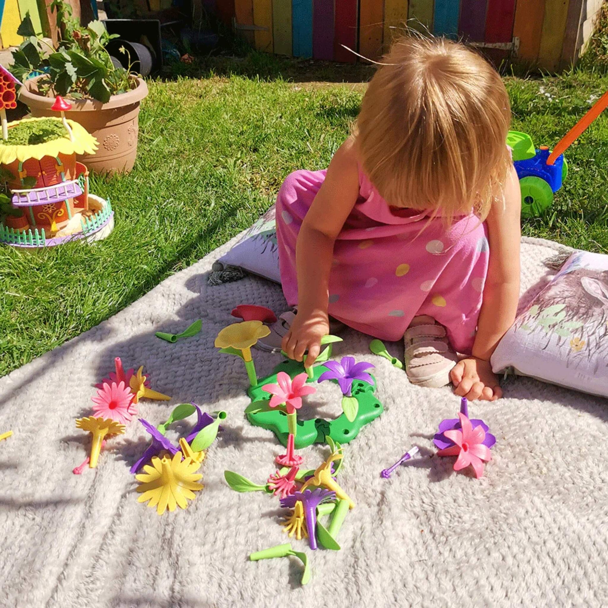 Toddler outdoors on a blanket, exploring loose flower pieces and arranging them into green interlocking bases on the grass.