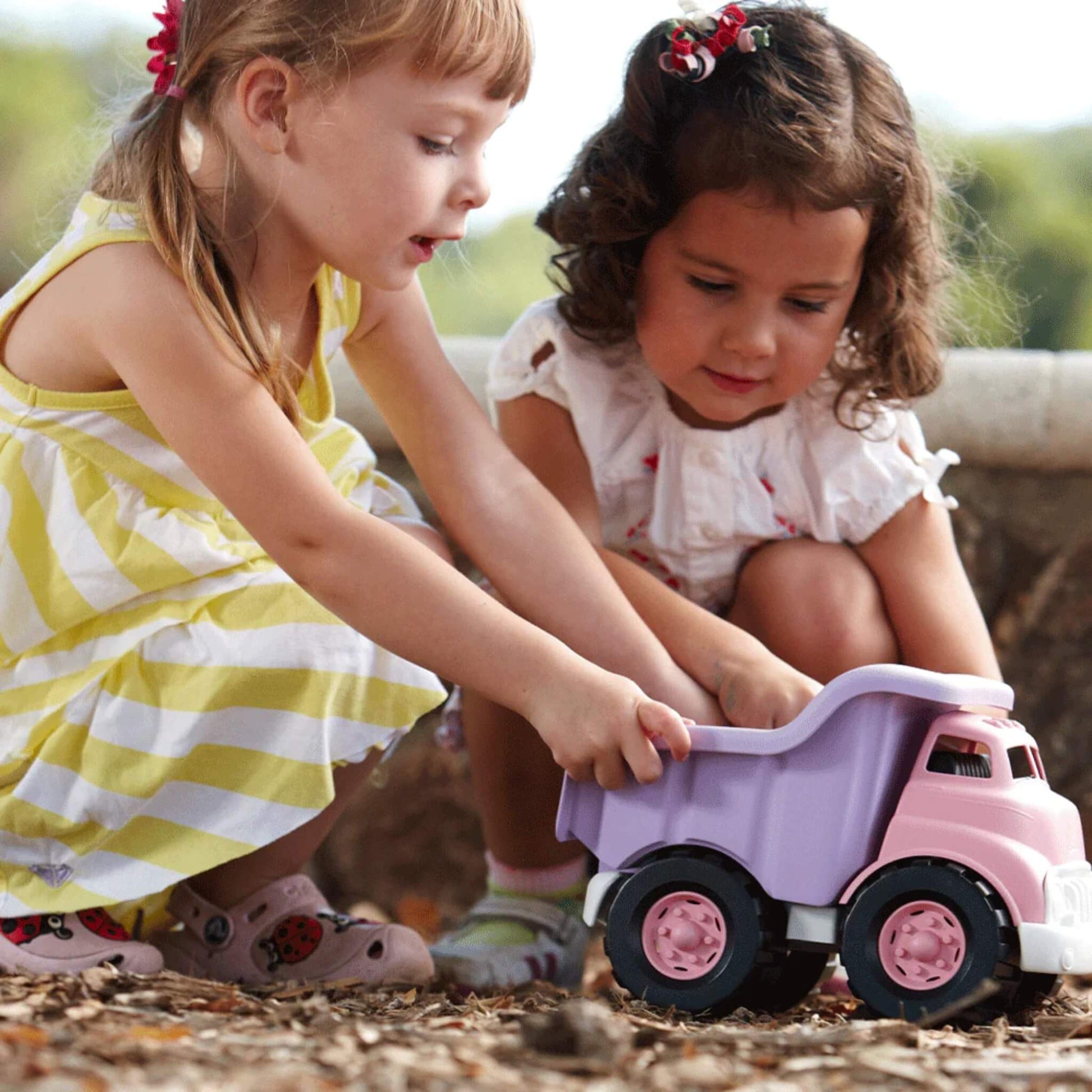 Two young girls crouched on a woodland path, playing together with the Green Toys pink and purple dump truck, both holding it as they explore the ground.