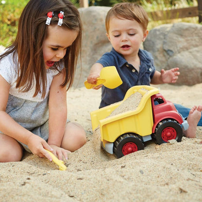 Young boy and girl playing in a sandy outdoor area, using a yellow shovel to fill the dump truck with sand.