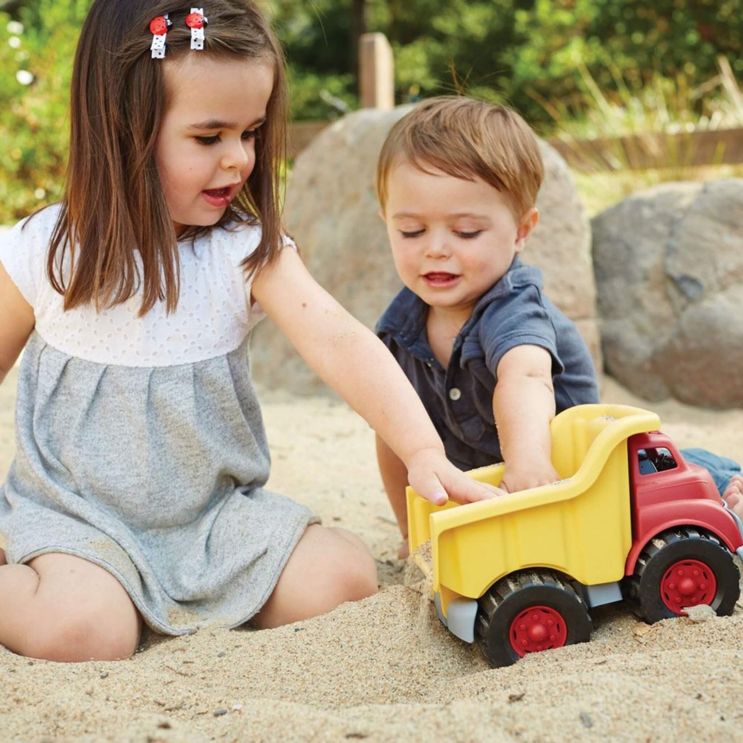 Two young children playing together in the sand, using their hands to load sand into the back of the dump truck.