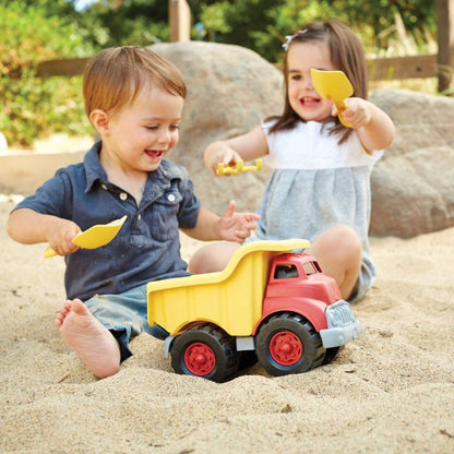 Two children sitting in a sandpit, smiling and holding toy shovels while playing with the dump truck positioned between them.