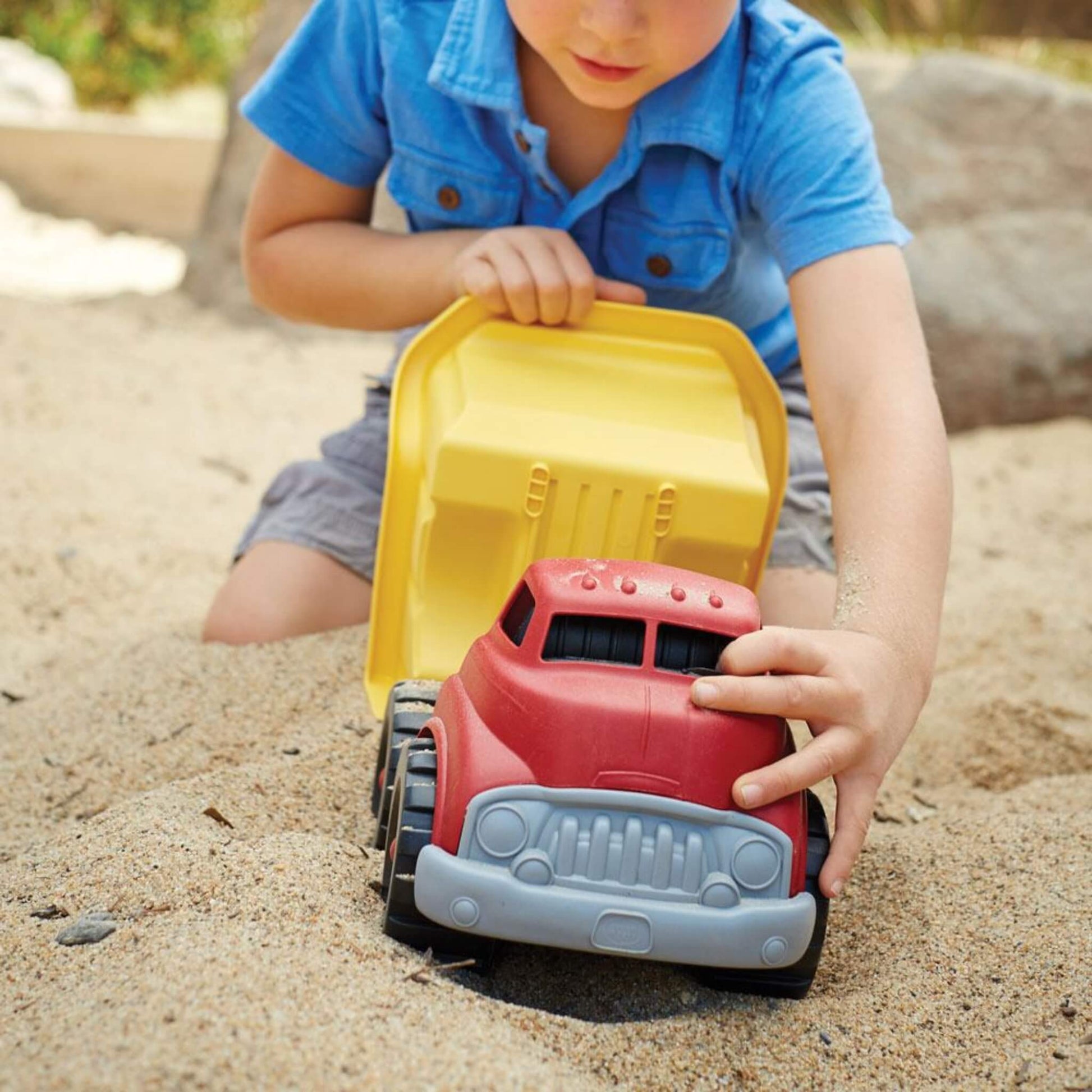Child in a blue shirt pushing the dump truck through sand, holding the front and the raised yellow dumper bed as they engage in play.
