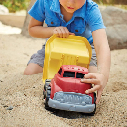 Child in a blue shirt pushing the dump truck through sand, holding the front and the raised yellow dumper bed as they engage in play.
