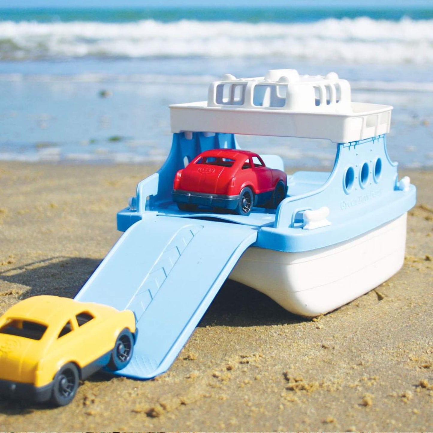 Ferry Boat set on a sandy beach with the red car on board and the yellow car approaching the ramp, facing the sea in the background.
