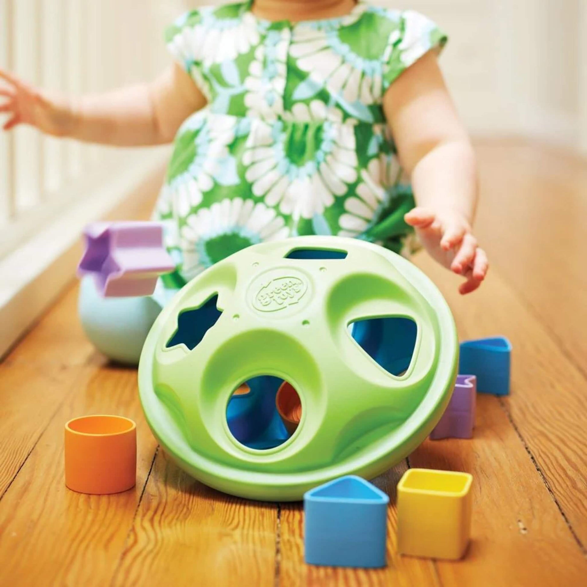 Close-up of baby in a green floral dress reaching toward the shape sorter surrounded by bright shapes on a wooden floor.