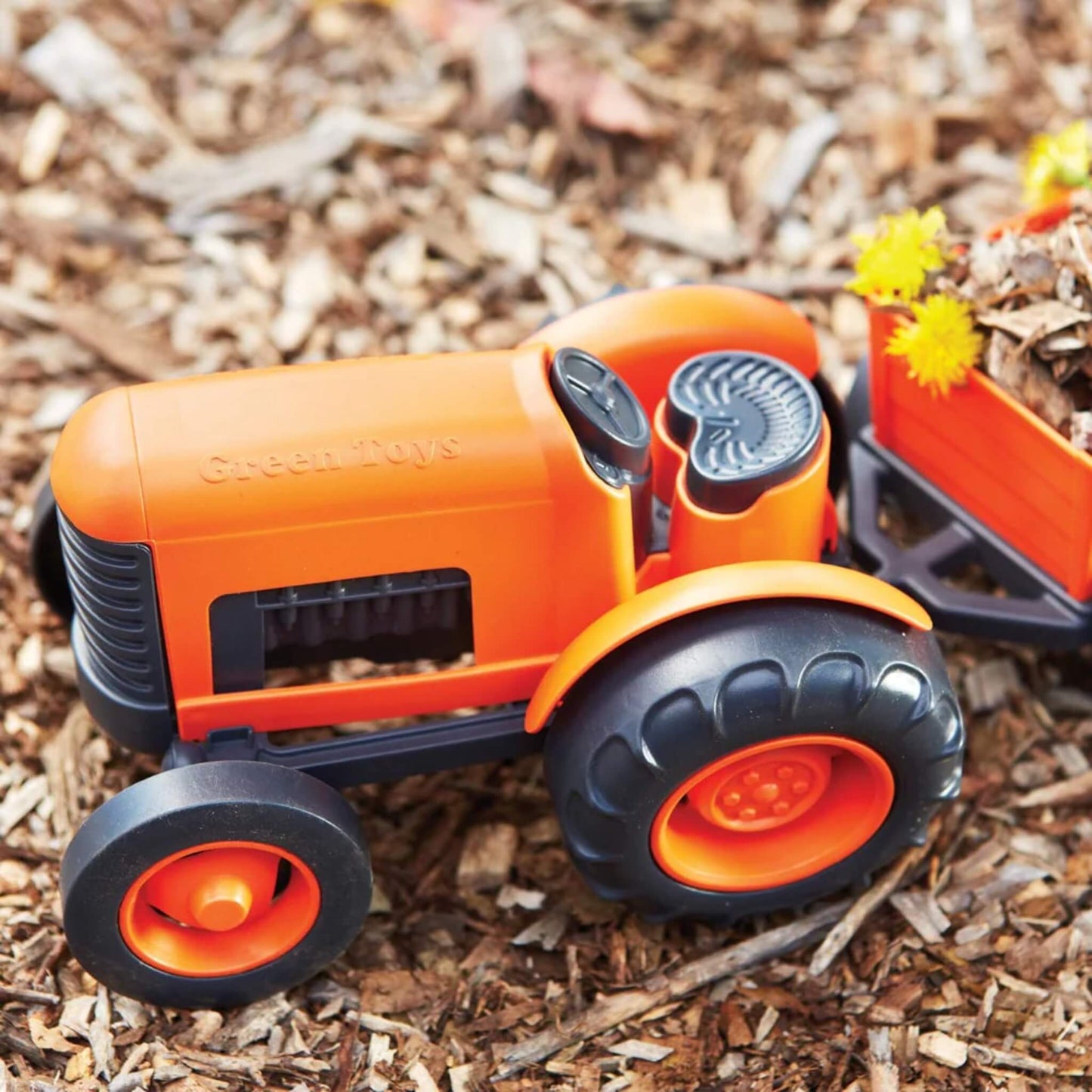 Toy tractor with rear trailer filled with bark and yellow flowers, placed outdoors on natural mulch surface.