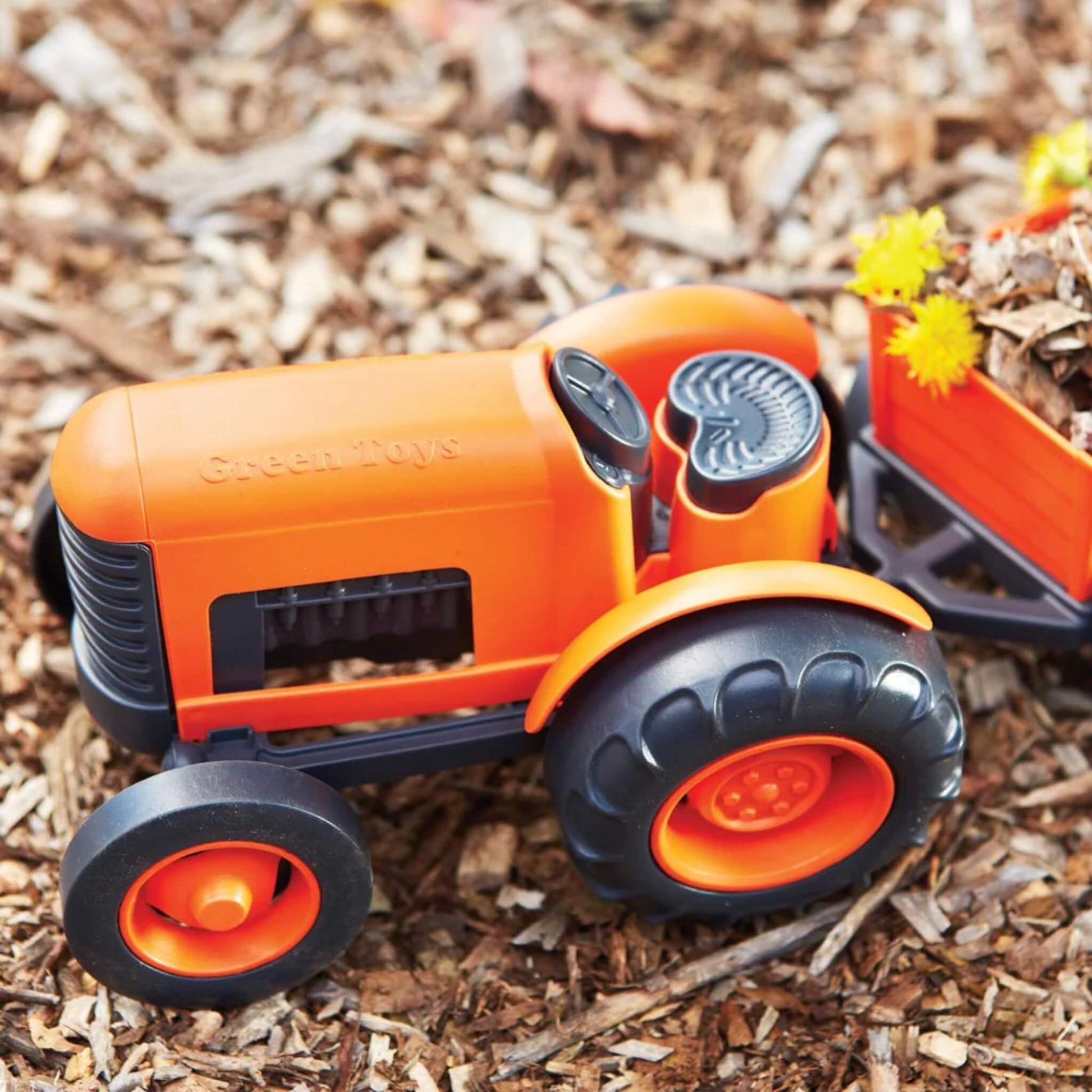 Toy tractor with rear trailer filled with bark and yellow flowers, placed outdoors on natural mulch surface.