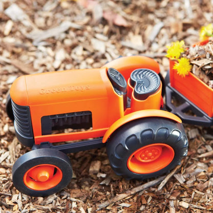 Toy tractor with rear trailer filled with bark and yellow flowers, placed outdoors on natural mulch surface.