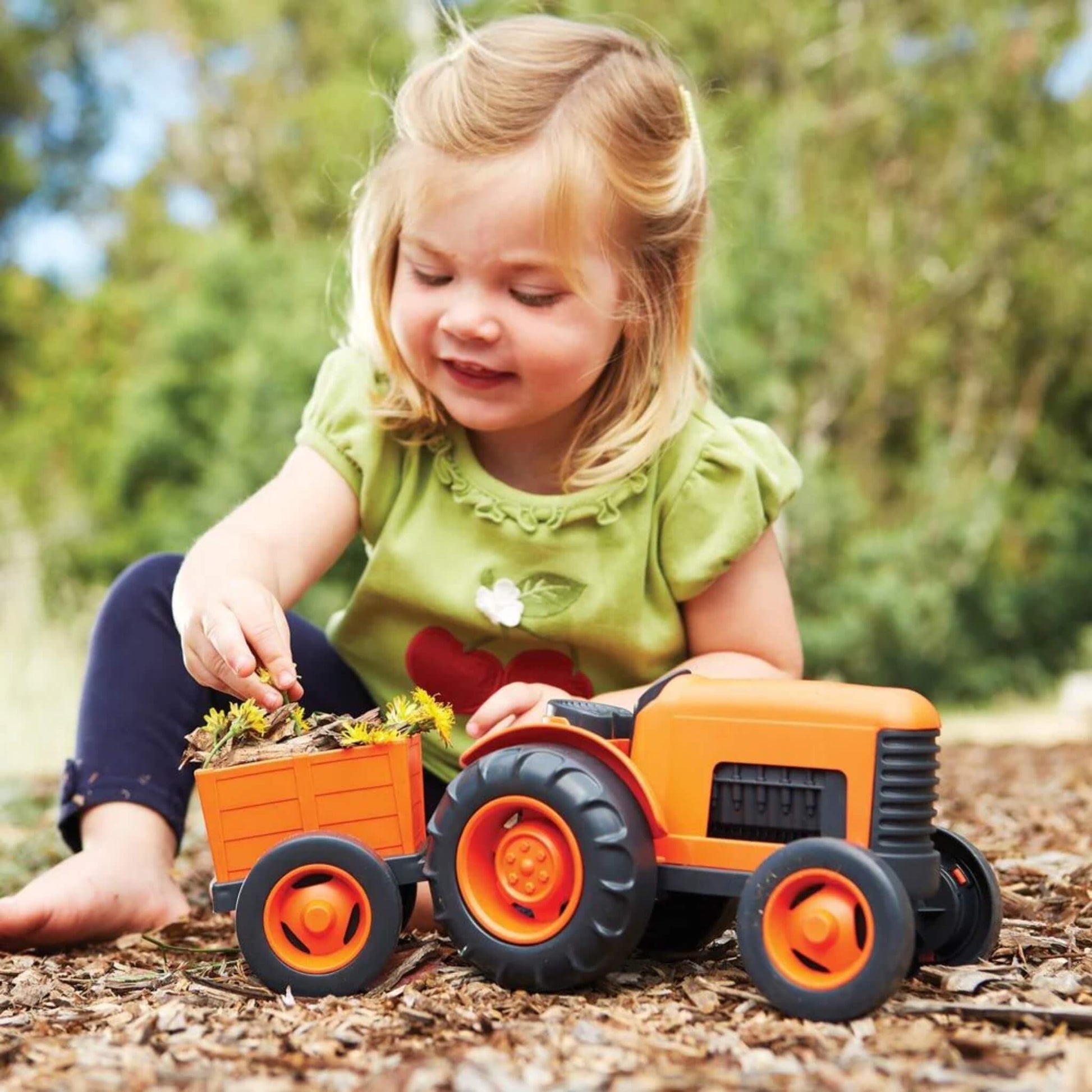 Young girl sitting barefoot on mulch while placing dandelions into the toy tractor’s trailer during outdoor play.