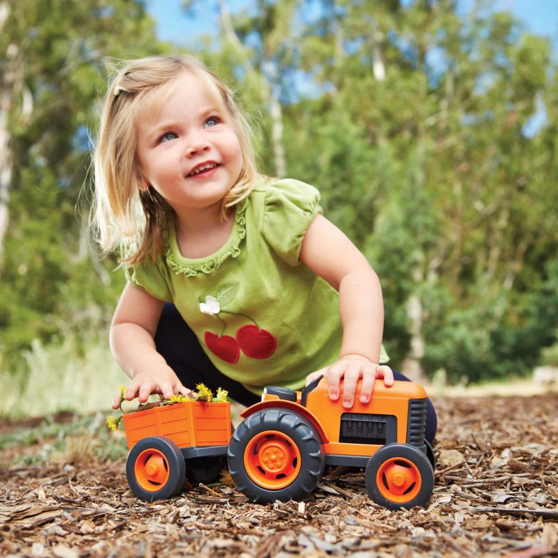 Smiling girl pushing the Green Toys tractor across a woodland area, holding the toy by its trailer.