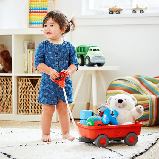 A toddler stands indoors holding the white cotton rope handle of the orange toy wagon, which is filled with toys including a plush bear and stacking cups.