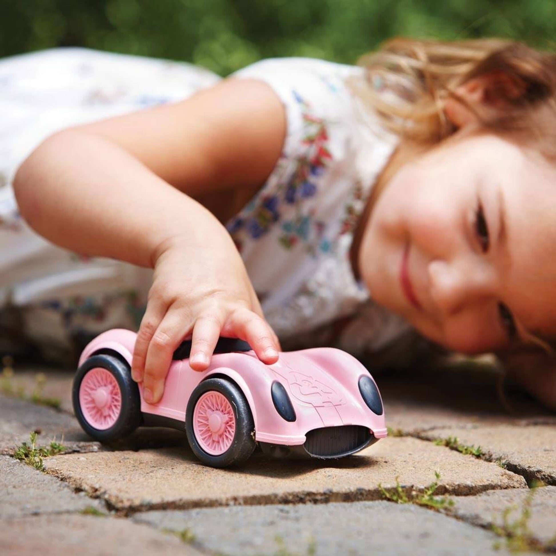 Young girl playing on a stone patio with a pink Green Toys Racing Car, smiling and pushing it along the ground with one hand.