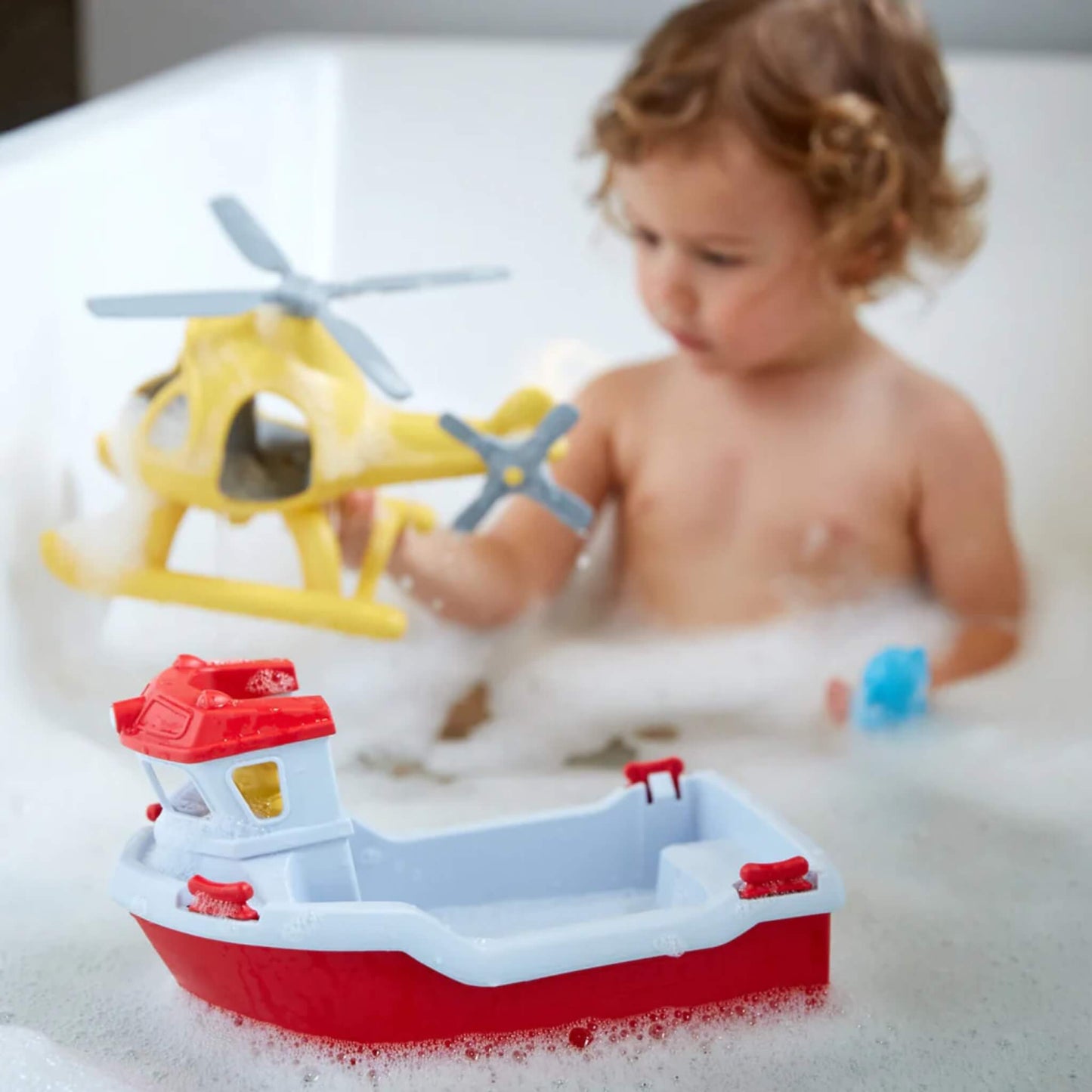 Young child plays in the bath with the Rescue Boat & Helicopter set; the red and white boat floats in the foreground while the child holds the yellow helicopter.