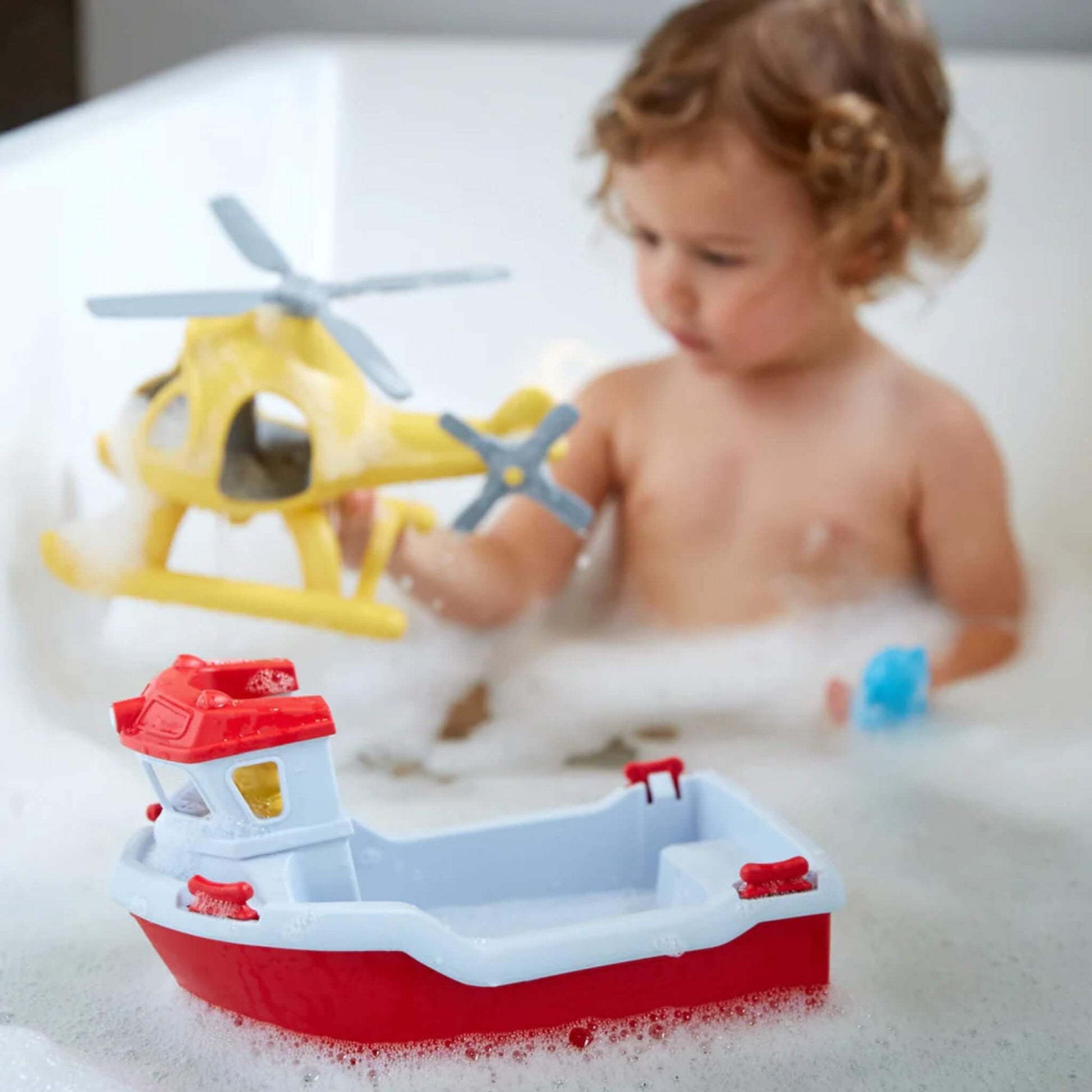 Young child plays in the bath with the Rescue Boat & Helicopter set; the red and white boat floats in the foreground while the child holds the yellow helicopter.