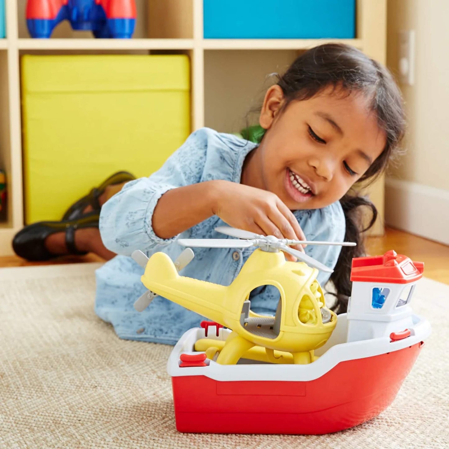 A smiling girl spins the blades of the yellow helicopter while it rests on the rescue boat, demonstrating indoor imaginative play.