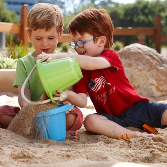 Two children playing in a sandpit—one boy holds the Green Toys green bucket while the other helps guide the sand into a blue bucket, both smiling and engaged in play.