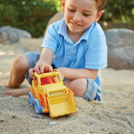 Young boy in a blue polo shirt playing with the scooper truck in a sandy outdoor area, pushing it forward with a focused expression.