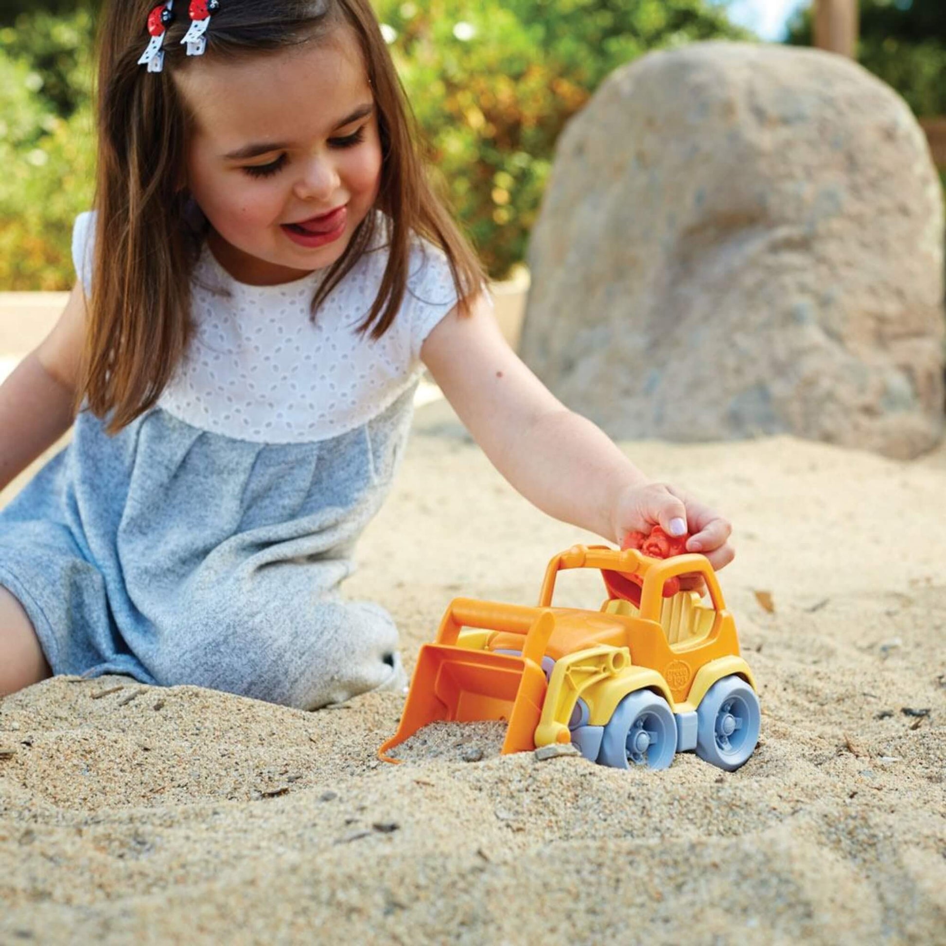 Young girl in a grey dress sitting in a sandpit, holding the scooper truck by the cab while gently pushing it along the sand.