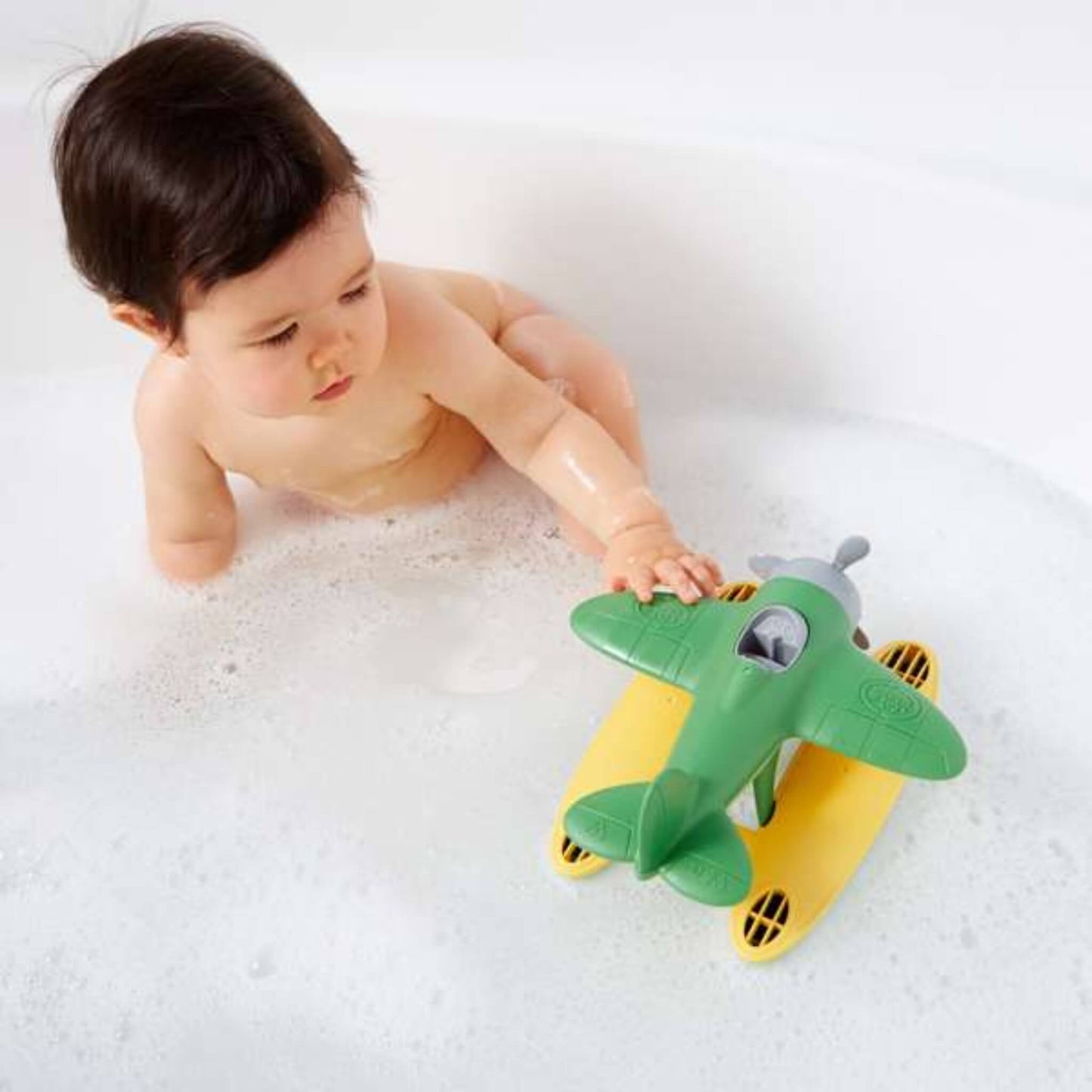 Baby sitting in a bubble bath reaching for the floating Green Toys Seaplane.
