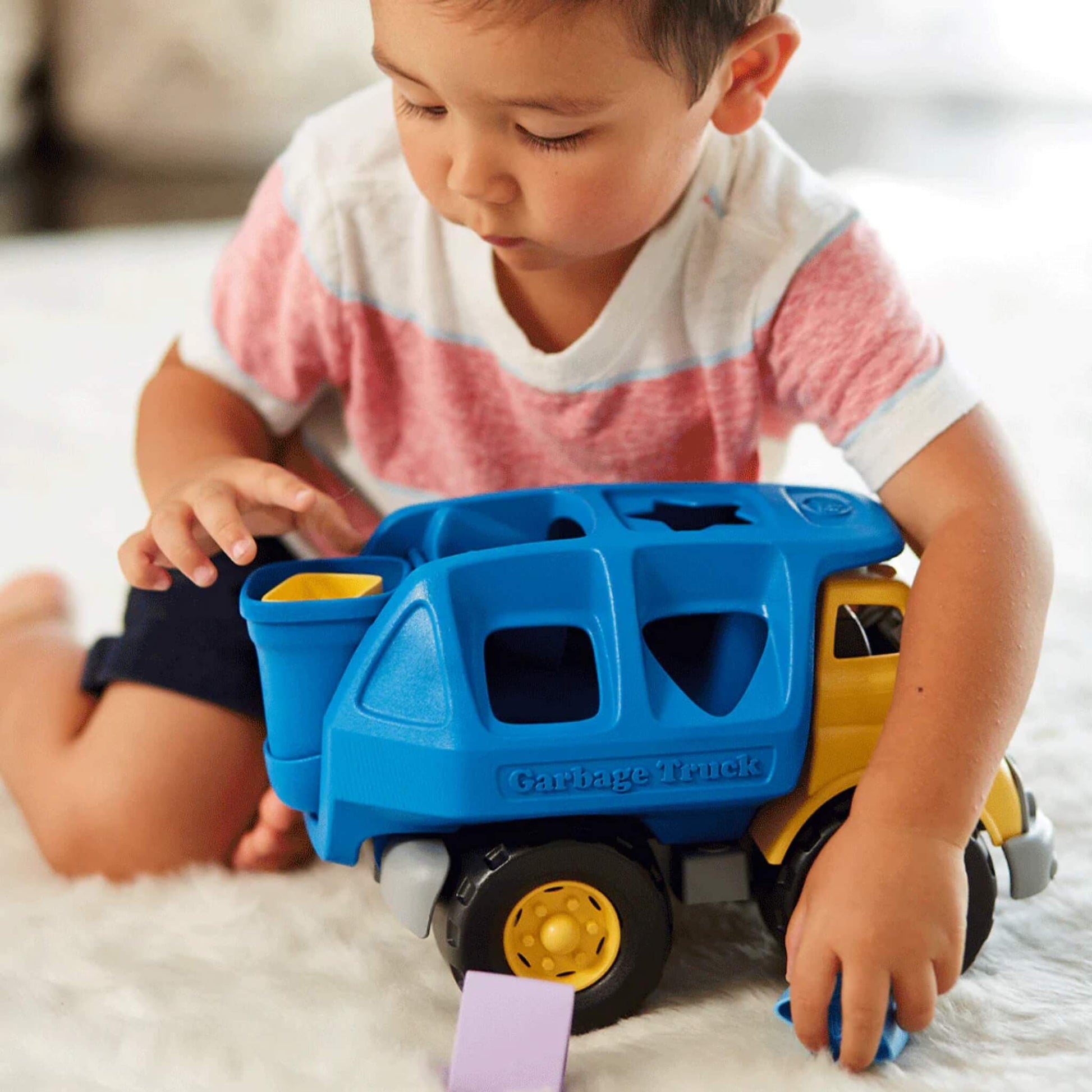 Toddler sitting on a white rug, focused on placing a yellow shape into the truck's rear container.