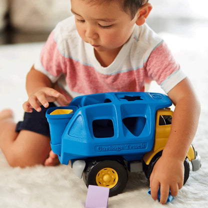 Toddler sitting on a white rug, focused on placing a yellow shape into the truck's rear container.