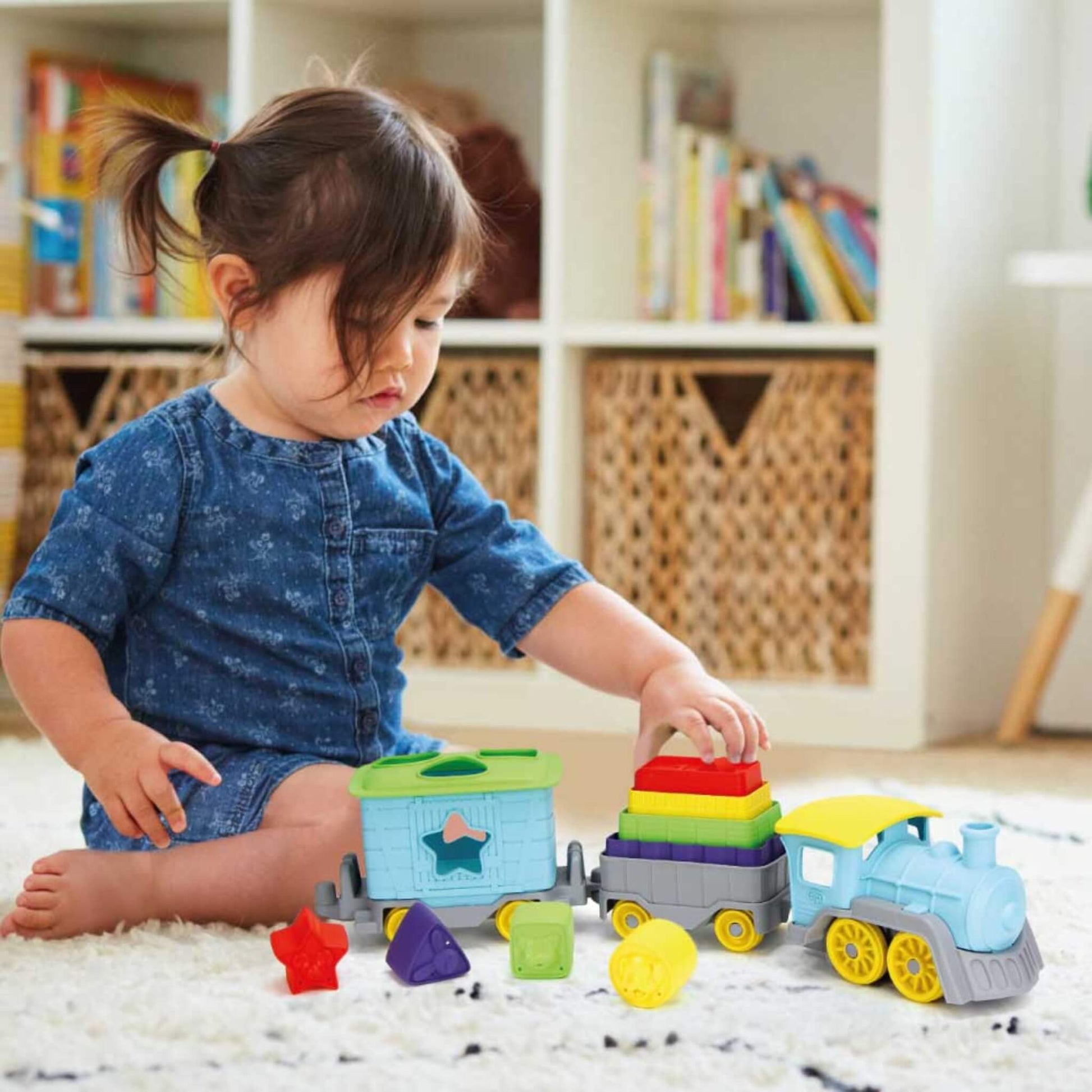 Toddler sitting on a rug placing stacking blocks onto the middle carriage of the train set, with colourful shapes scattered around.