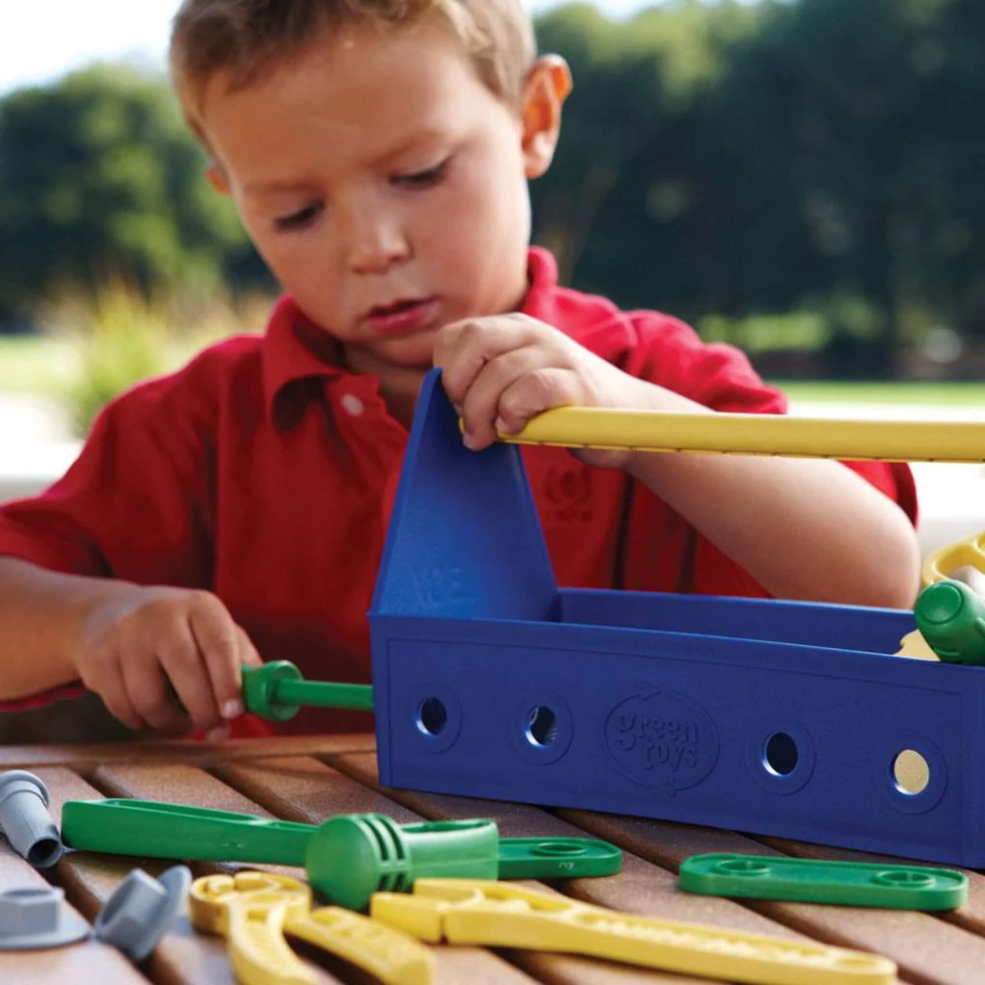 A young child in a red shirt playing outdoors with the blue toolbox, using a toy screwdriver to fasten a plastic bolt.