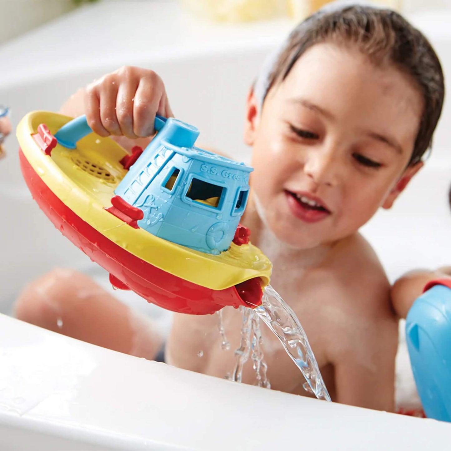 A young child playing in the bath, pouring water from the spout of the tugboat with a happy expression.
