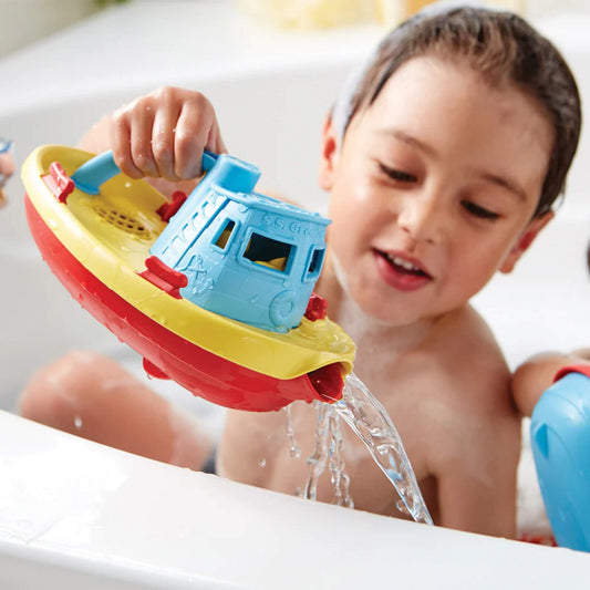 A young child playing in the bath, pouring water from the spout of the tugboat with a happy expression.