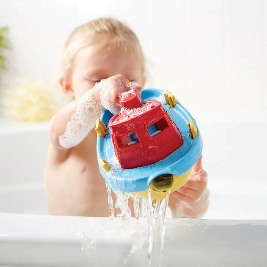 A toddler holding the Green Toys Tugboat above a bathtub, pouring water through the front spout while covered in soap suds.