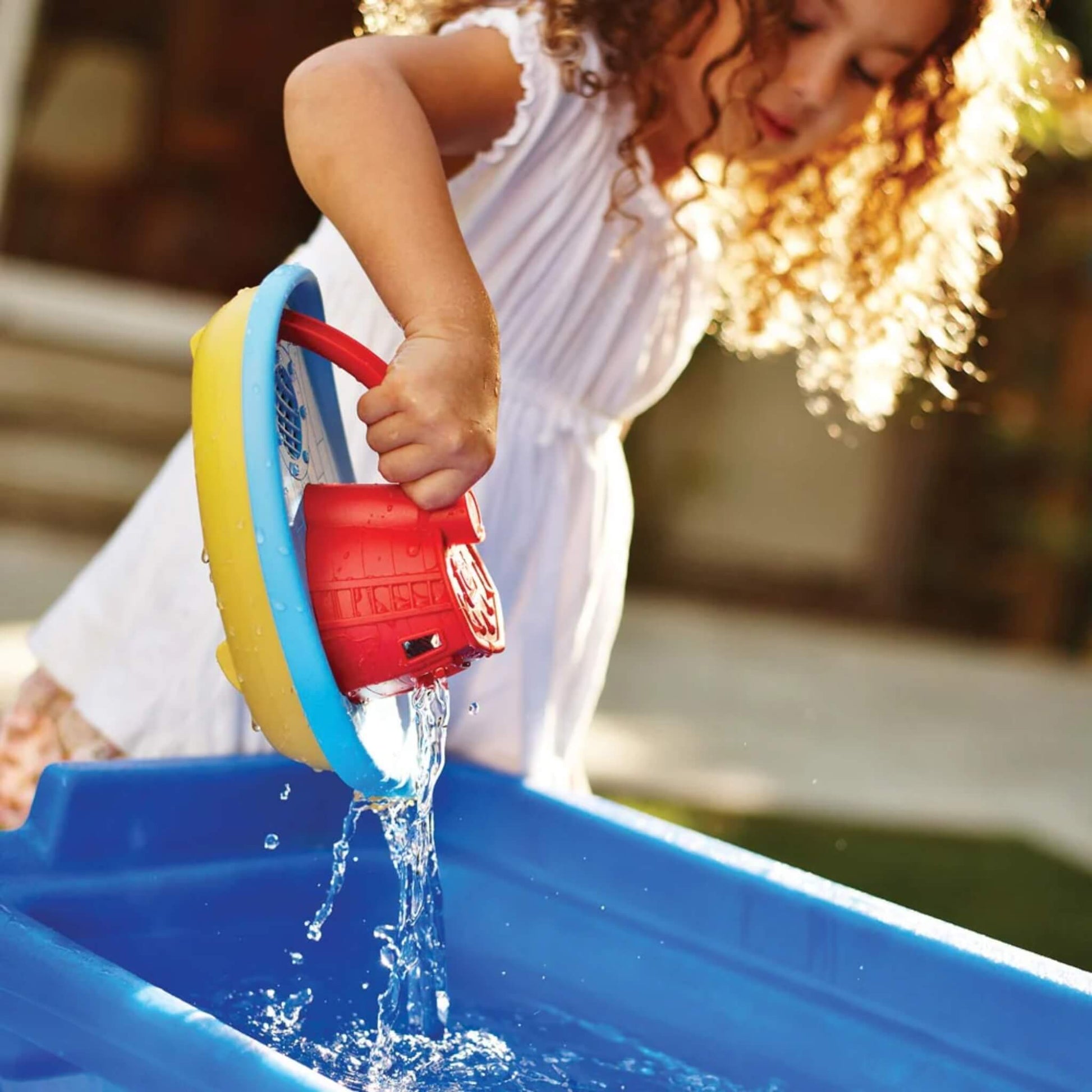 A young girl outdoors pours water from the tugboat into a blue container, showing how the toy works outside the bath too.