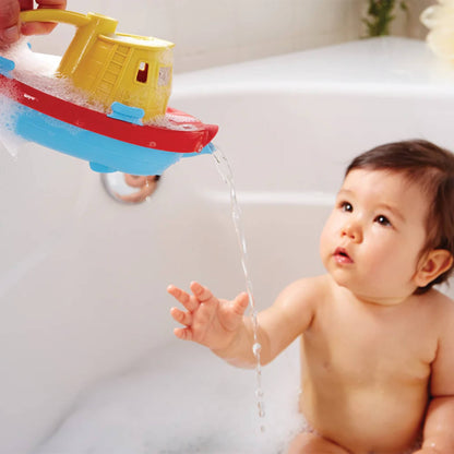 A baby sitting in the bath reaches towards the tugboat as an adult pours water gently from its spout.
