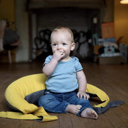 Baby sitting upright on the floor supported by a crescent-shaped feeding pillow used as a prop cushion.