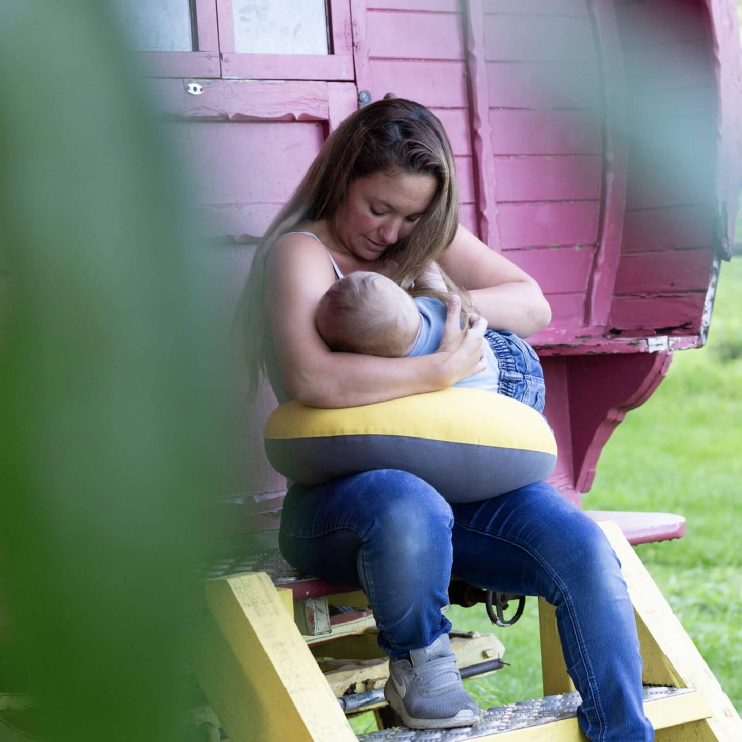 Mother sitting outdoors on wooden steps, holding her baby with a crescent-shaped feeding pillow wrapped around her waist.