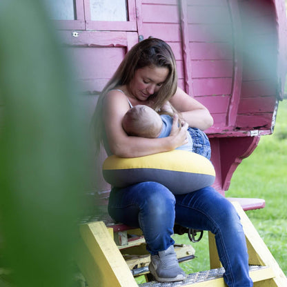Mother sitting outdoors on wooden steps, holding her baby with a crescent-shaped feeding pillow wrapped around her waist.