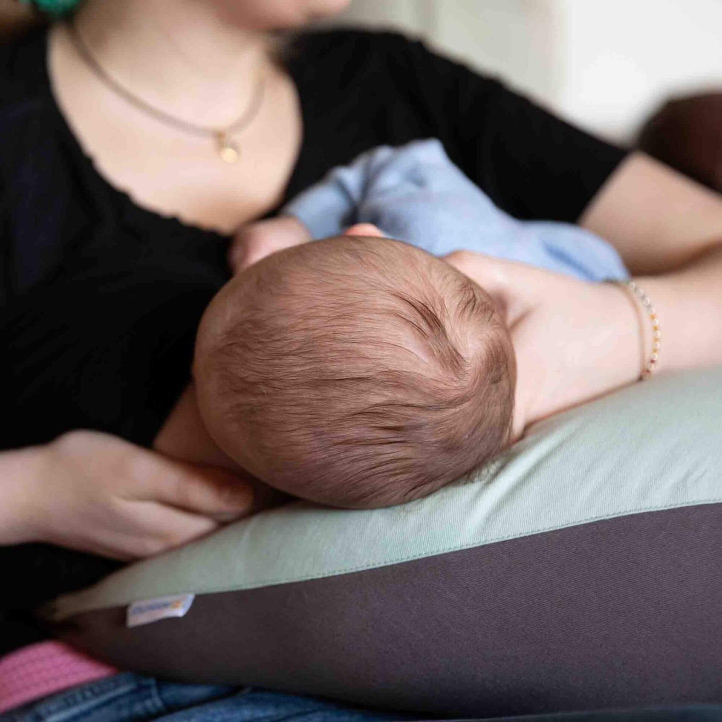 Mother holding her baby in a breastfeeding position with a crescent-shaped sage green and charcoal feeding pillow across her lap.