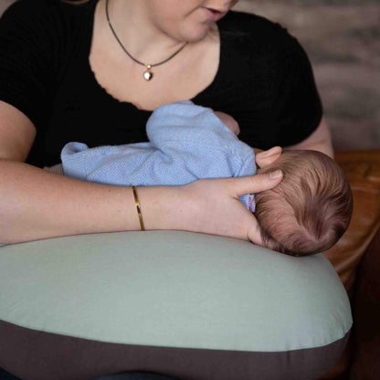 Baby lying across a crescent-shaped sage green feeding pillow, supported in a feeding position by their mother’s arms.
