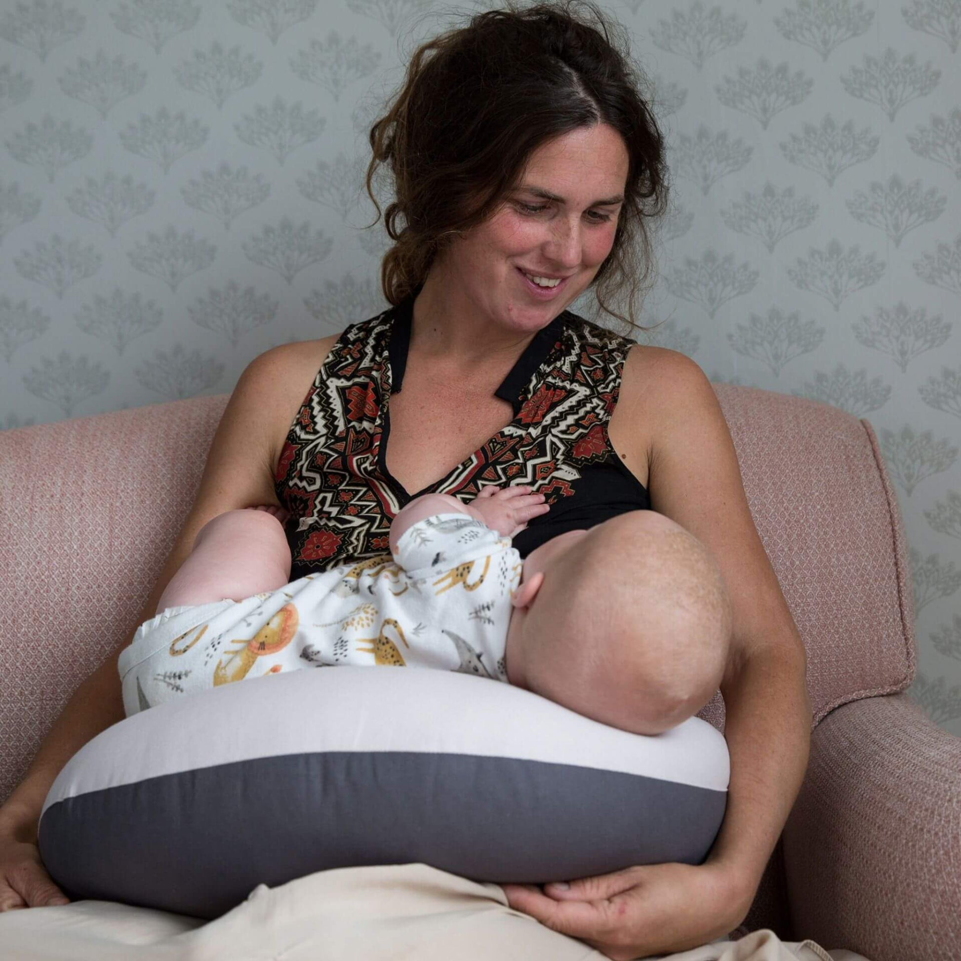 Mother seated in an armchair breastfeeding her baby using a dusky pink feeding pillow across her lap, with the pillow supporting the infant at chest height.