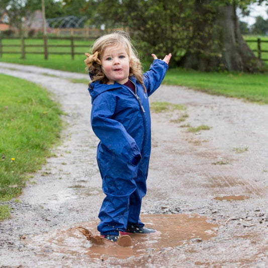 Toddler wearing a blue waterproof all-in-one suit while splashing in a muddy puddle.