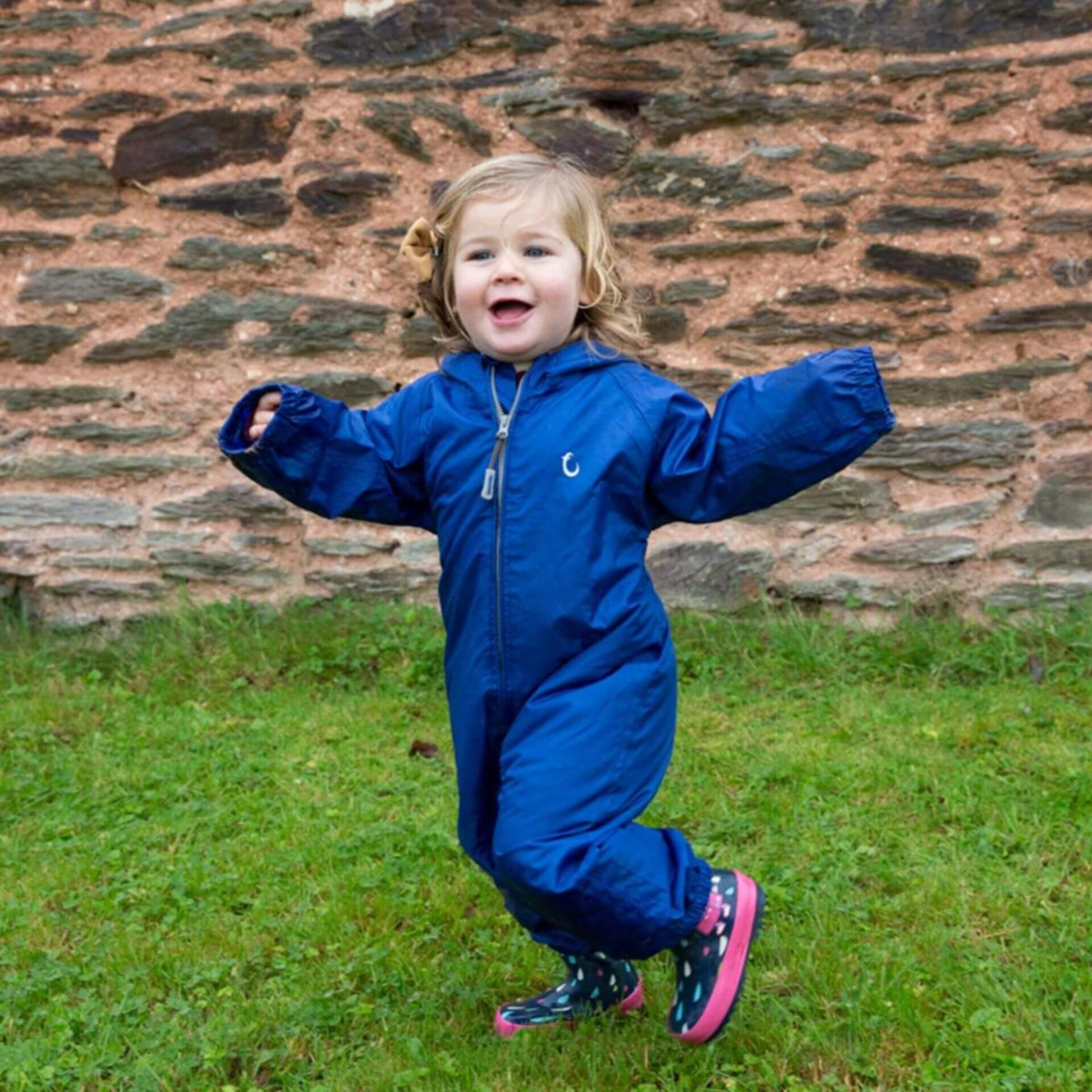 Young child playing outside in a blue waterproof suit standing near a stone wall.