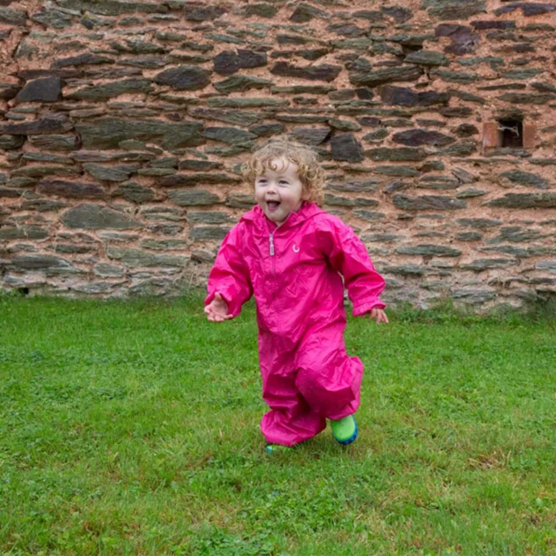 Toddler running happily on grass while wearing a bright pink waterproof Packasuit, with a stone wall in the background.