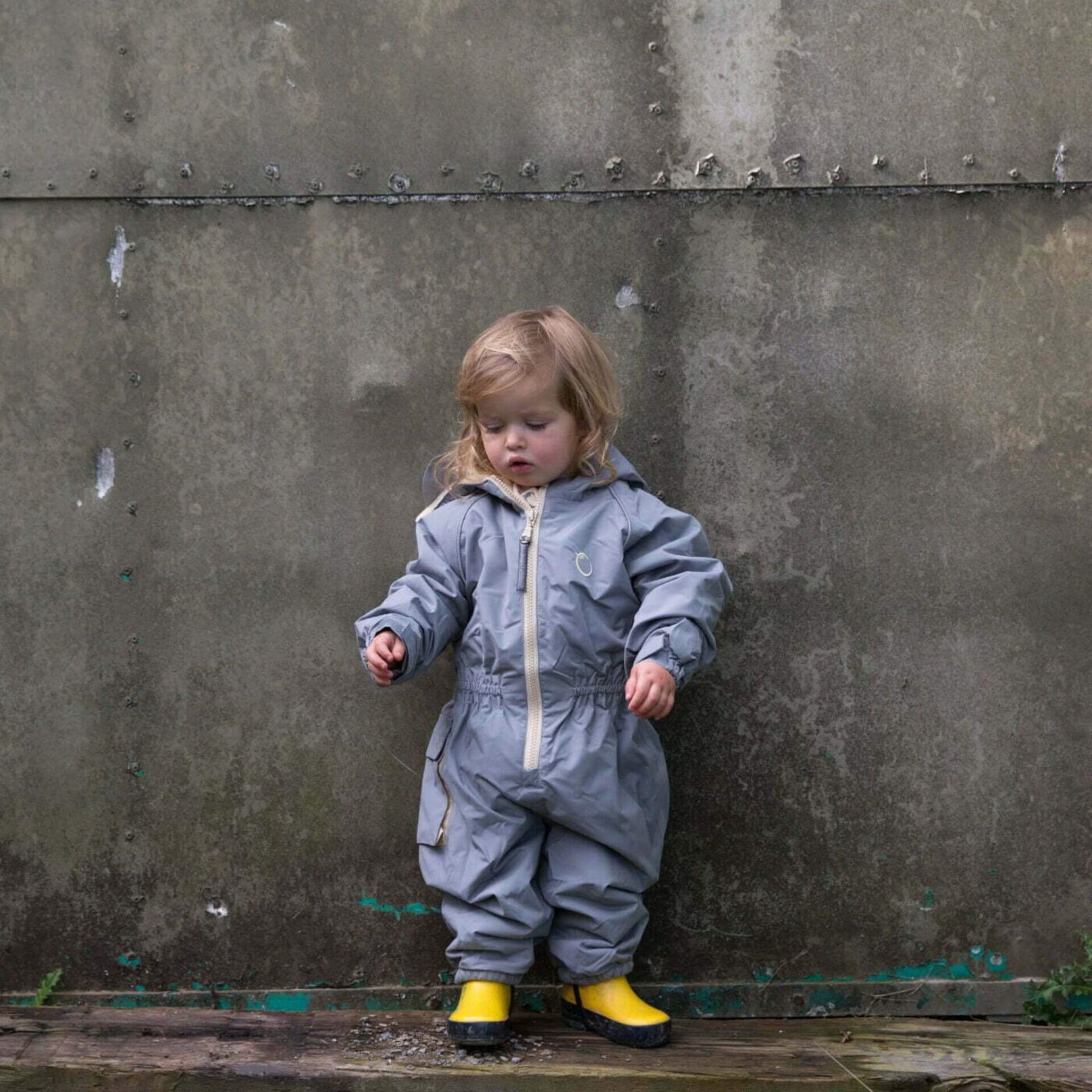 Toddler in a grey waterproof fleece lined suit with yellow boots, standing against a concrete wall outdoors.