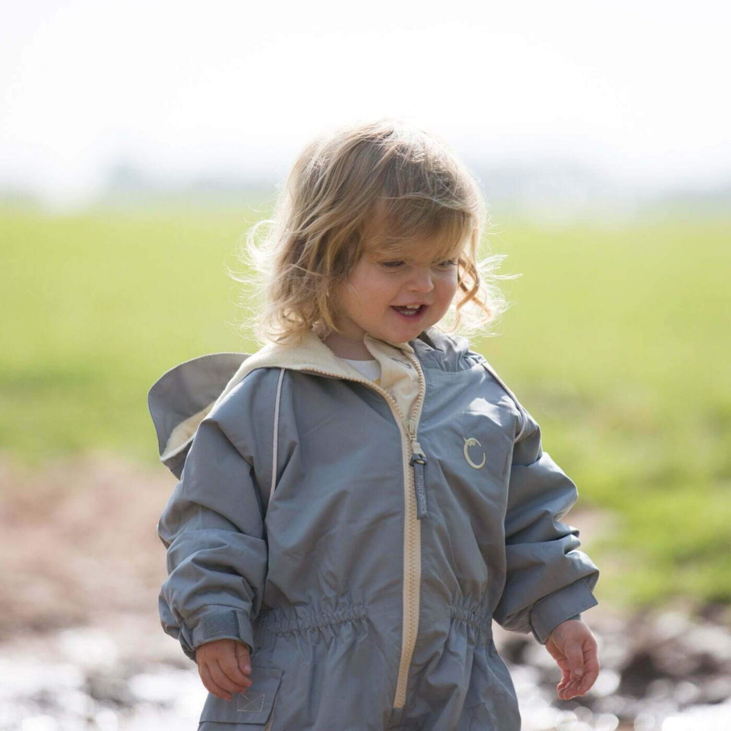 Close-up of toddler smiling while wearing a grey fleece lined waterproof suit with beige zip and hood, standing on a muddy path.