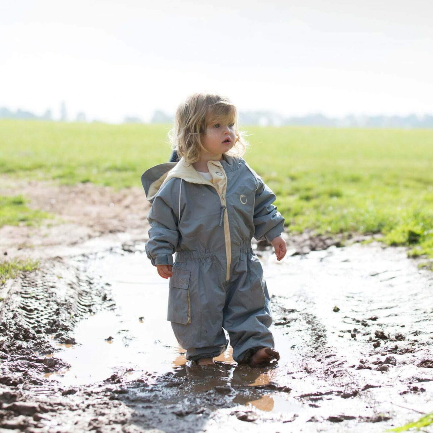 Toddler standing in a muddy puddle wearing a grey waterproof fleece lined suit with hood and beige zip, playing outdoors on a bright day.