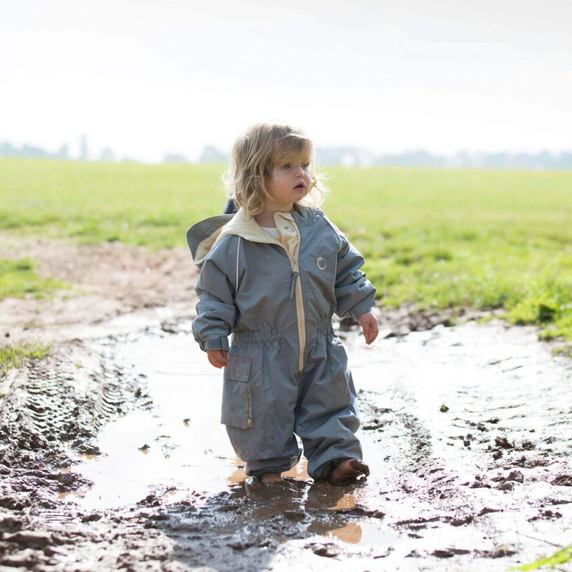 Toddler standing in a muddy puddle wearing a grey waterproof fleece lined suit with hood and beige zip, playing outdoors on a bright day.