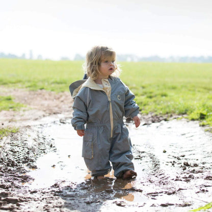 Toddler standing in a muddy puddle wearing a grey waterproof fleece lined suit with hood and beige zip, playing outdoors on a bright day.